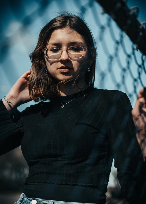 Jeune femme portant des lunettes, en haut noir, posant près d'un grillage